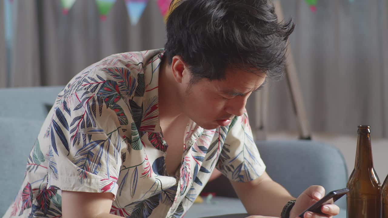술에 취한 아시아인 남자가 소파에 앉아서 집에서 파티를 한 후 스마트폰을 연주하고 있다.