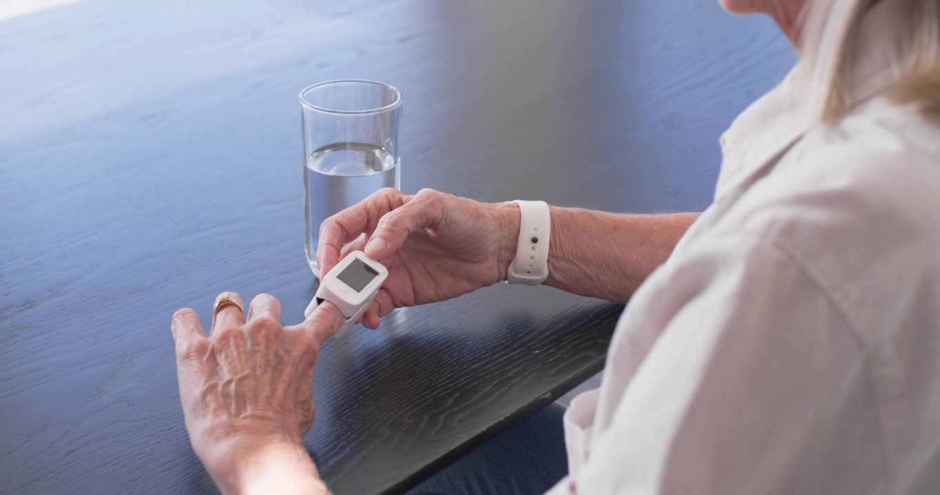Senior woman sitting at home table, putting oximeter on left finger, monitoring oxygen, copy space