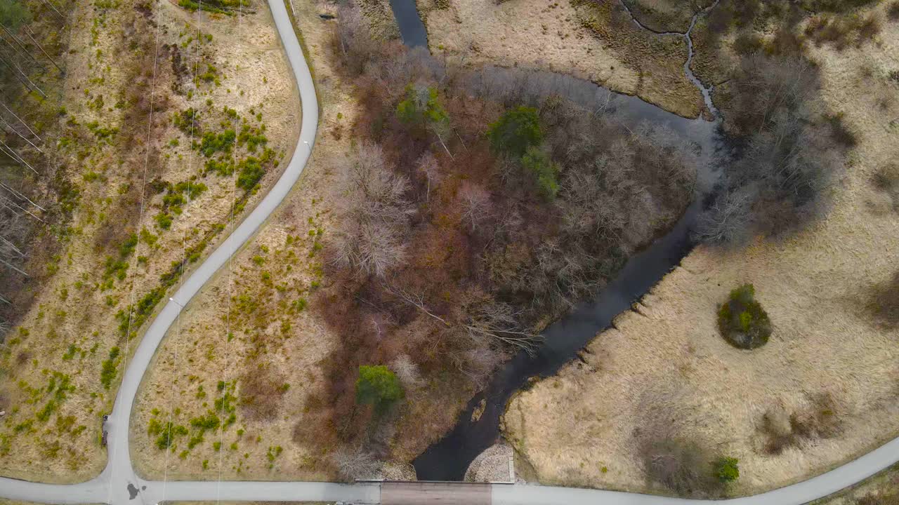 Top down aerial footage flying over and revealing Pääsküla dark brown colored river during spring or autumn time when nature and grass is brown and yellow. Paved roads and a wooden bridge visible also
