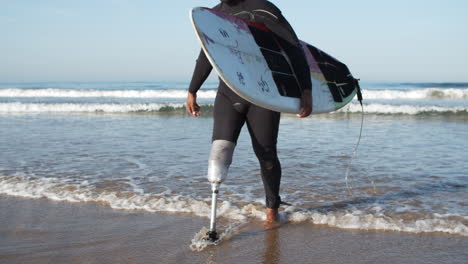 vista frontal de un surfista serio con una pierna biónica saliendo del océano sosteniendo una tabla de surf bajo el brazo