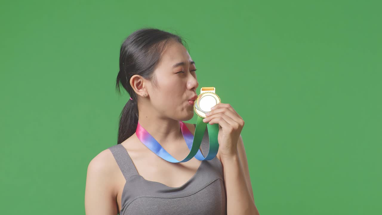 Close Up Of Asian Woman Kissing A Gold Medal In Her Hand Being Proud Winning As The First Winner On Green Screen Background In The Studio