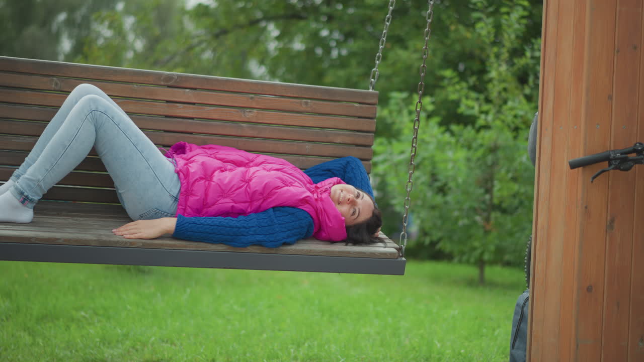 Kaukasische Frau liegt auf einer hölzernen Schaukelbank, trägt eine leuchtend rosa Weste und einen blauen Pullover, barfuß in hellen Jeans, entspannt, Knie angewinkelt, den Blick zum Himmel über den Rahmen eines abgelegenen grünen Rasens gerichtet