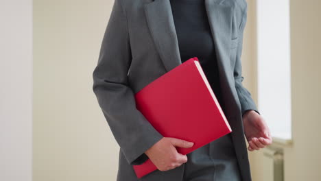 businesswoman in gray suit holding red folder standing indoors ready for meeting or presentation with confident body language and professional attire in office or corporate environment
