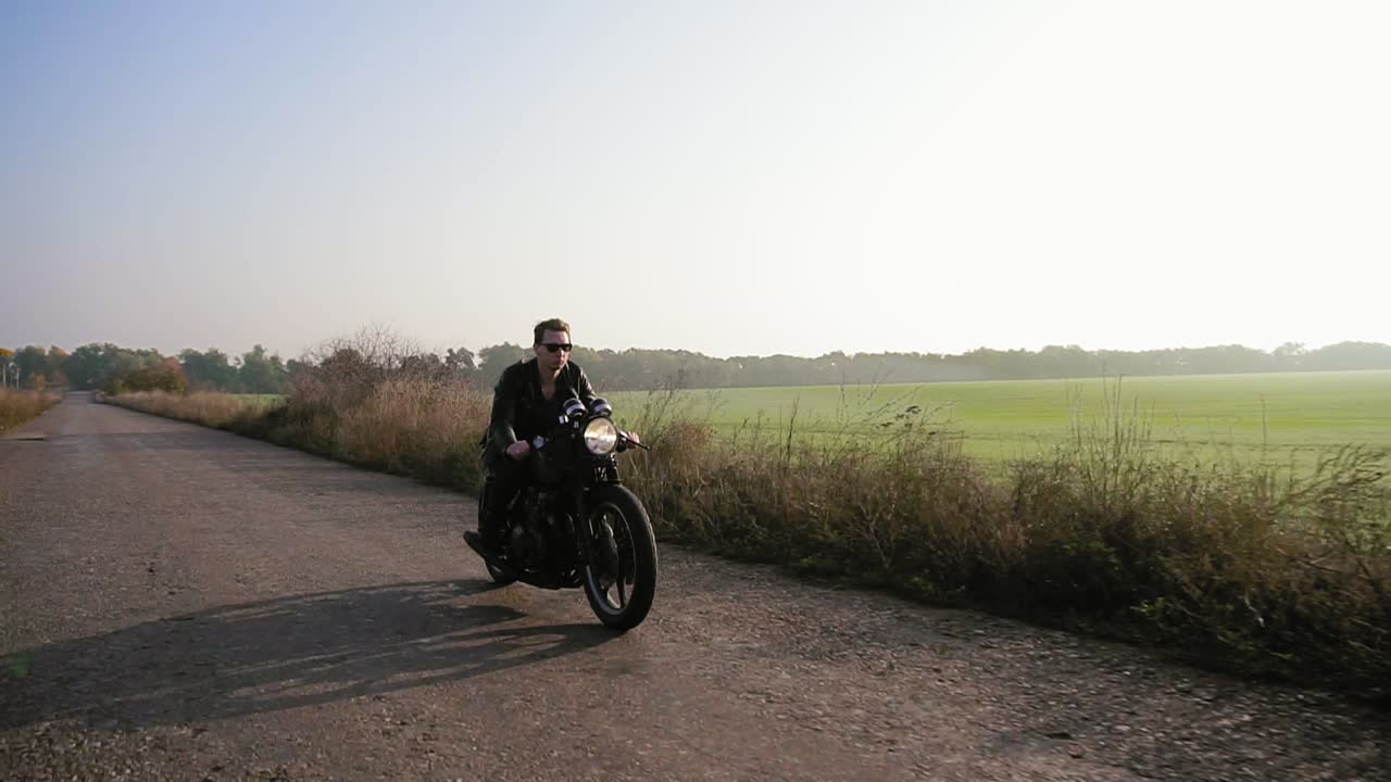 ⁇  안경과 가죽 재 ⁇ 을 입은 세련된 시원한 젊은 남자가 가을  ⁇ 은 날 아스팔트 도로에서 오토바이를 타고 있습니다.
