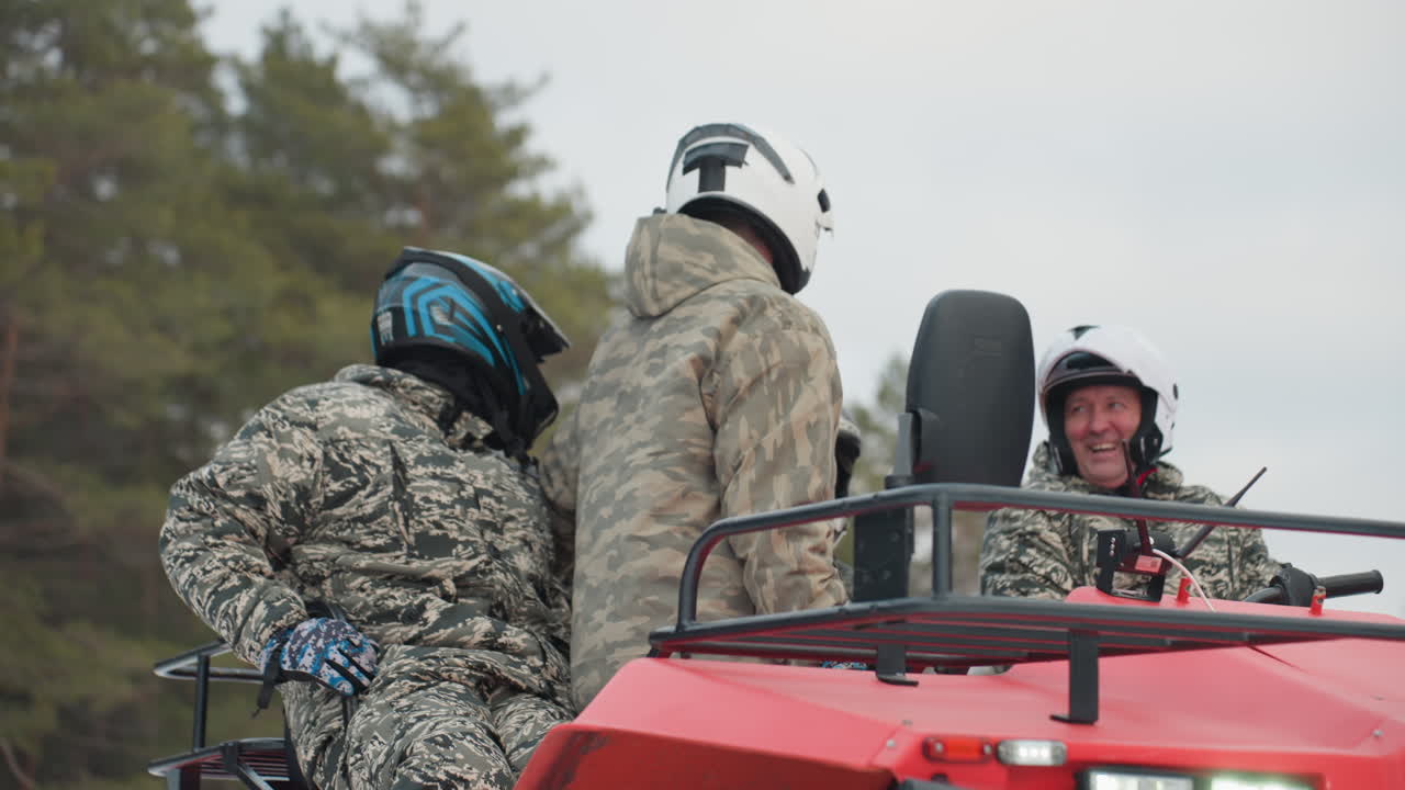 Three helmeted riders prepare beside atv in snowy pine forest, camouflage jackets and visors, close conversational briefing and equipment check, guide instructs passengers, focused expressions convey