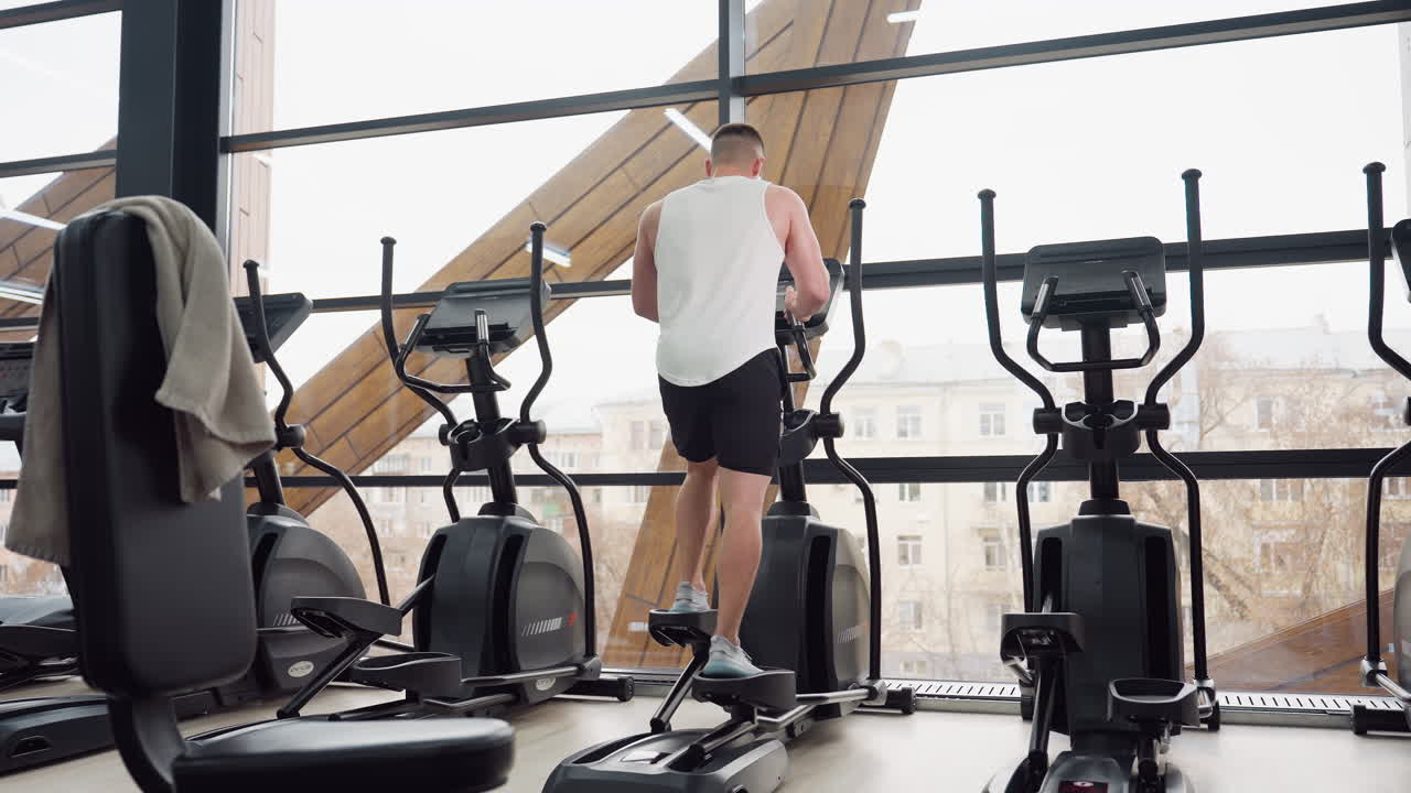 Athlete exercising on elliptical trainer in modern athletic club with towel draped over chair, stepping on pedals by slanted windows overlooking street, adjusting console and maintaining posture