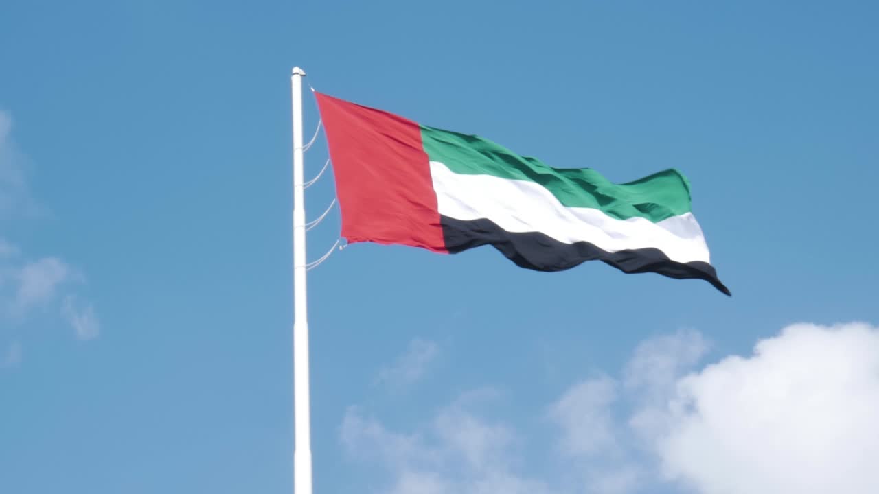 Slow-motion shot of the flag of the United Arab Emirates, waving in the wind, against a bright blue sky with some clouds.