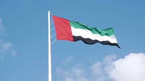 slow-motion shot of the flag of the united arab emirates, waving in the wind, against a bright blue sky with some clouds.