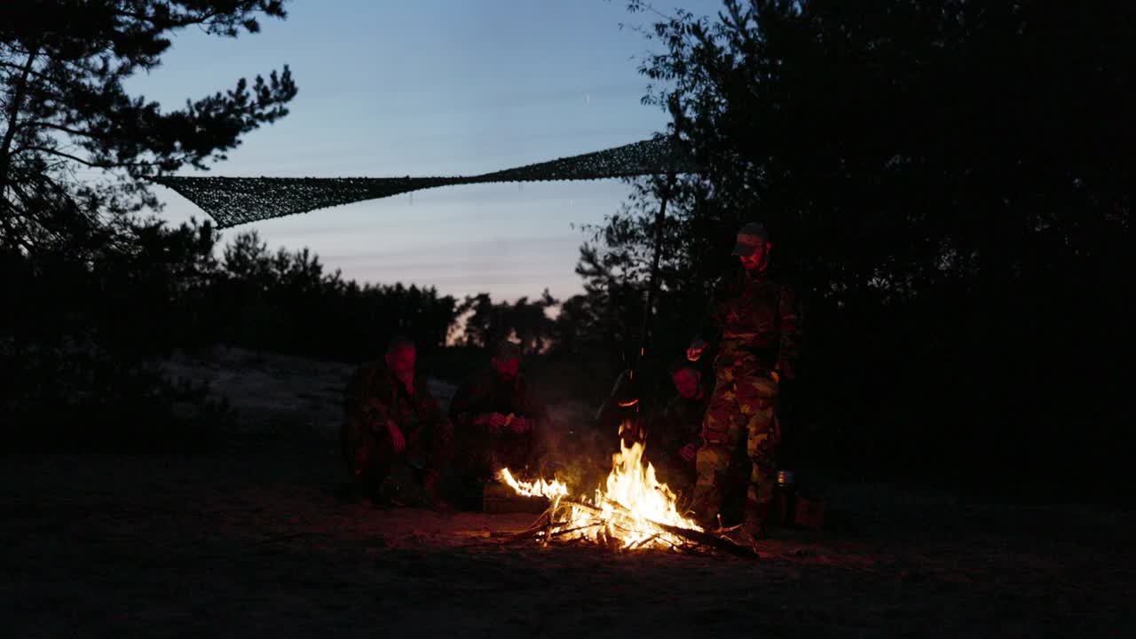 위장복을 입은 한 무리의 남자들이 밤에 들판에 있는 언덕 위에 앉아 불타는 캠프불 에서 몸을 따뜻하게 하고, 저녁 식사로 소시지를 고, 남성적인 외출, 일 후 휴식을 취하고, 봉사하고, 사냥하고 있습니다.
