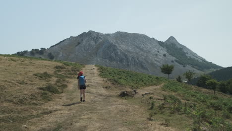 Vista-Trasera-De-Una-Mujer-Joven-Caminando-Hacia-La-Montaña-De-Piedra-Caliza-De-Anboto-En-El-País-Vasco-Occidental-De-España-Mientras-Lleva-Una-Bolsa-Azul