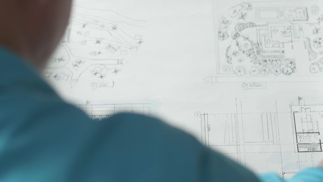 Close Up Of A Man Engineer Looking At Blueprint While Working At The Office, Over Shoulder