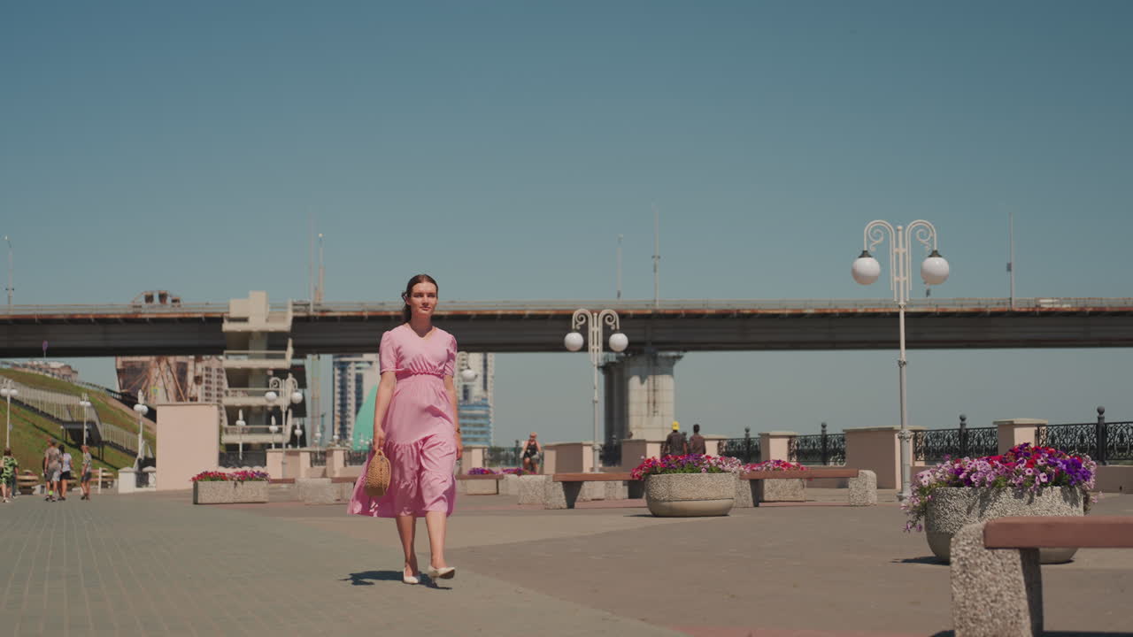 Mulher caminhando pelo calçadão à beira do rio sob um céu azul claro, com uma ponte ao fundo e vasos de concreto cheios de flores vibrantes, vestido casual de verão, ritmo cinematográfico lento, brisa suave