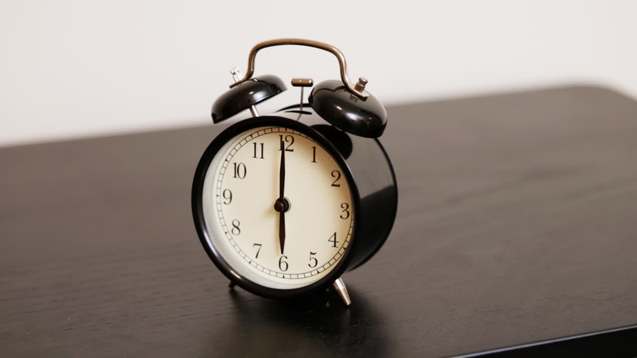 A black twin-bell analog alarm clock sits still, displaying 6:00 AM. The classic bedside timepiece represents early mornings, punctuality, and the calm before the alarm rings.