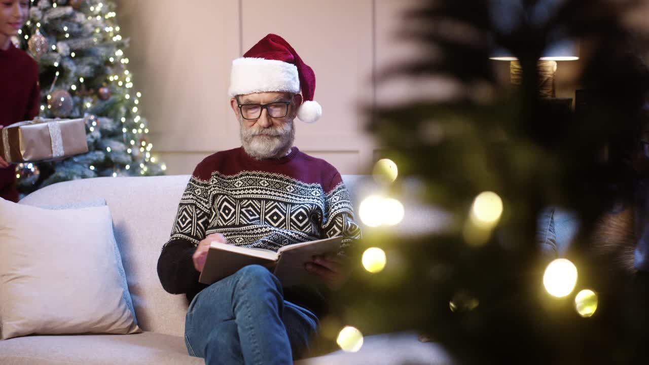un alegre abuelo de pelo gris con sombrero de santa que recibe regalos de navidad envueltos de sus nietos mientras se sienta en una casa decorada cerca de un árbol de navidad resplandeciente y un libro de lectura