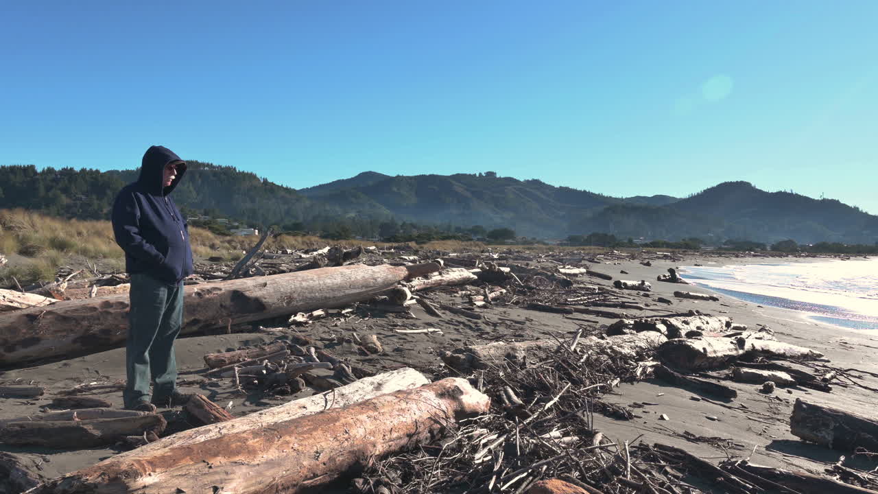 hombre con chaqueta con capucha parado sobre troncos en gold beach, costa de oregón - plano amplio y estático