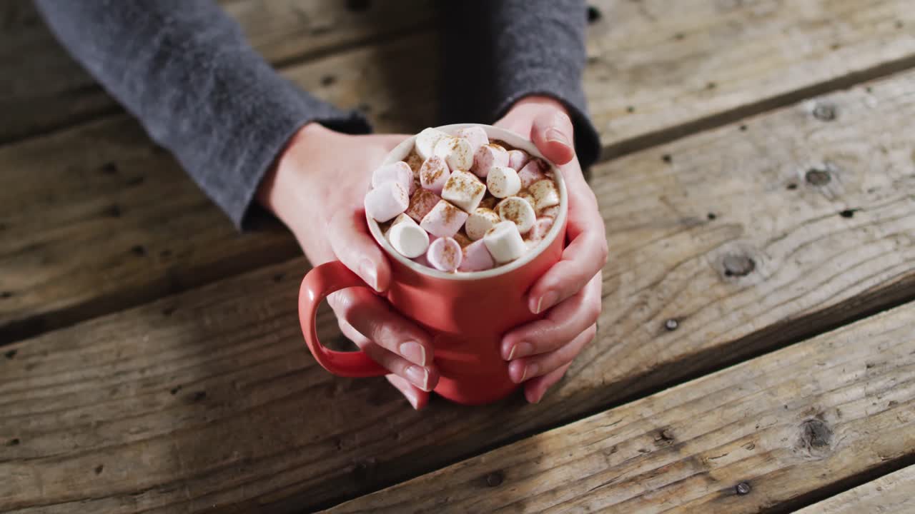vista de cerca de las manos sosteniendo un chocolate caliente con malvaviscos contra la superficie de madera