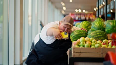 Ein-Supermarktangestellter-In-Einem-Weißen-T-Shirt-Und-Einer-Schwarzen-Schürze-Mit-Einer-Braunen-Mütze-Macht-Ein-Selfie-Und-Verzieht-Das-Gesicht-Neben-Grünen-Wassermelonen-Im-Supermarkt