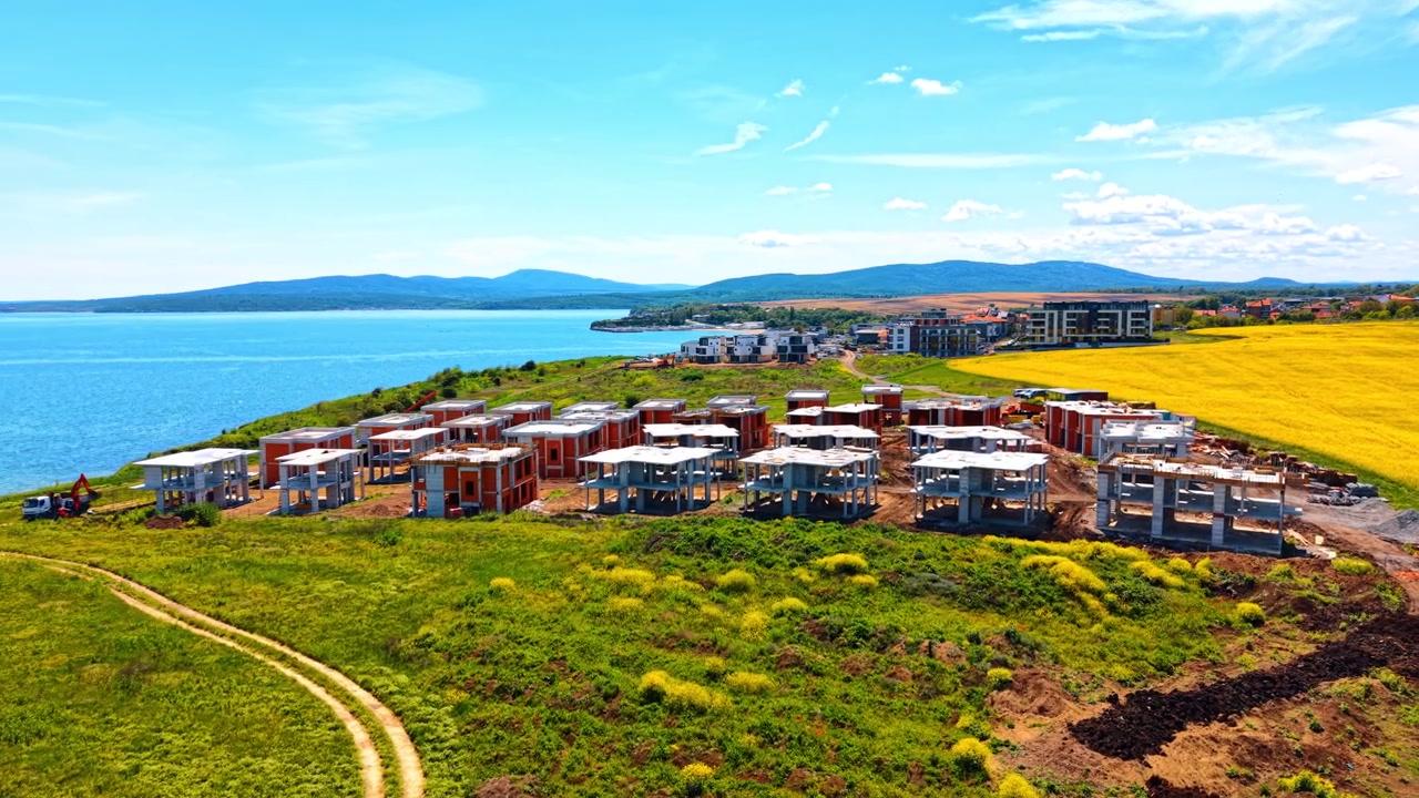 Sarı bir tarlanın yakınındaki kıyı konut projesi gelişiminin havadan görünümü. Çiçek açan bir kolza tarlasının bitişiğindeki yeşil bir kıyıda bir kır evi köyü inşaatının drone çekimi.