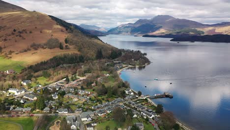 ángulo de retroceso aéreo alrededor del pueblo de luss en loch lomond sobre el agua para revelar el pueblo a principios de la primavera