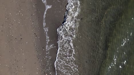 Una-Vista-De-Arriba-Hacia-Abajo-Sobre-Una-Playa-Vacía-Mientras-Las-Olas-Chocan-Suavemente-Contra-La-Orilla-En-Cámara-Lenta