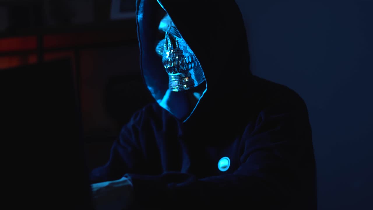 Masked Figure in Hoodie Typing in a Dark Room – Depicting Hacking, Encryption Theft, Cybersecurity Threats and Network Attacks