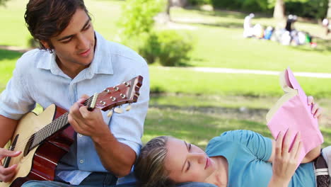 el hombre practica la guitarra mientras la mujer descansa en su pierna y lee.