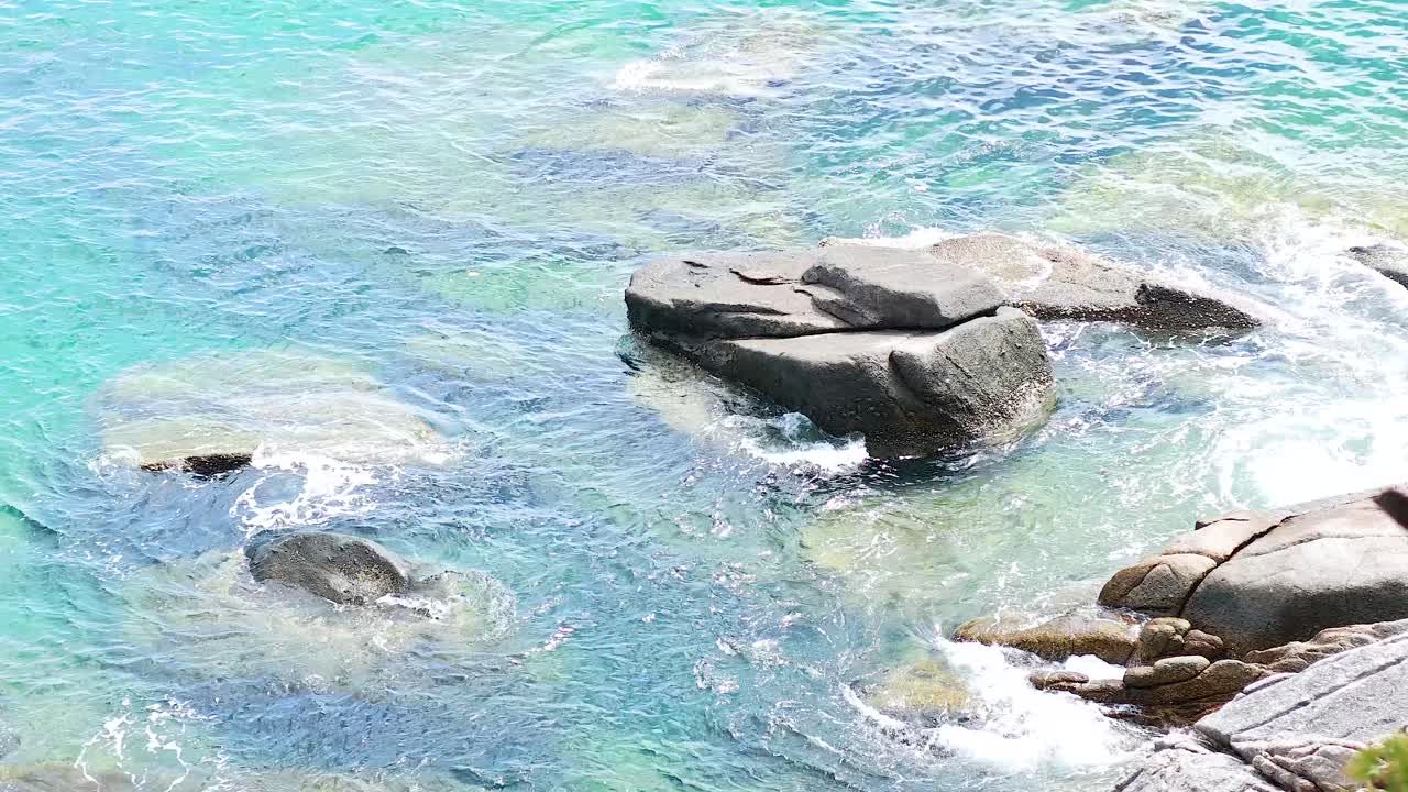Turquoise waves gently crash against coastal rocks under bright sunlight, creating a serene and dynamic seascape