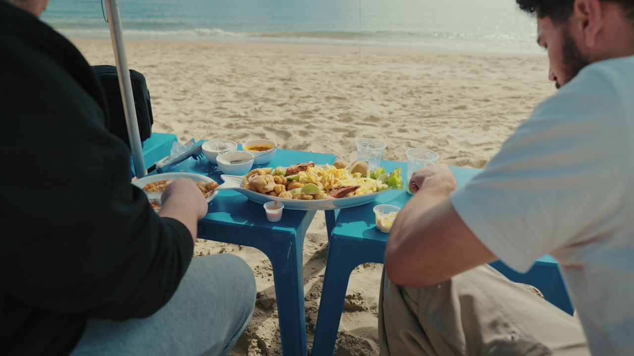 Two men enjoy fresh seafood at a blue table set directly on the sand, savoring Brazilian beach cuisine by the sea with a scenic oceanfront view