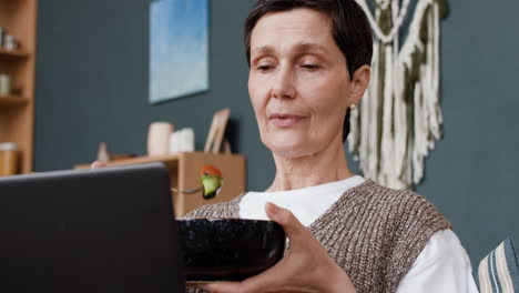 senior woman eating healthy meal while using laptop at home