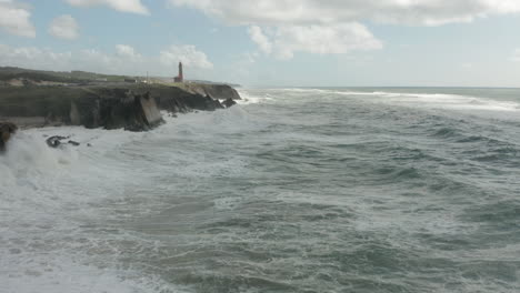 wild sea waves forming and splashing near rocky cliffs with a tall lighthouse in the background