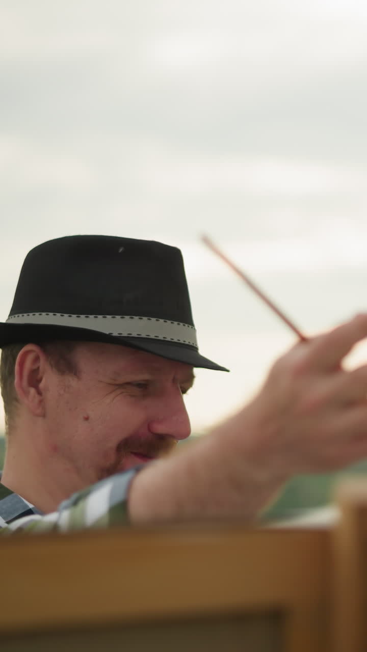 모자와 체크색 셔츠를 입은 화가가  드레스와 모자를 입은 여자가 웃기며 잔디 잎으로 코를 만지며 야외에서 가운 순간을 나누는 것처럼 그의 작업에 집중하고 있습니다.