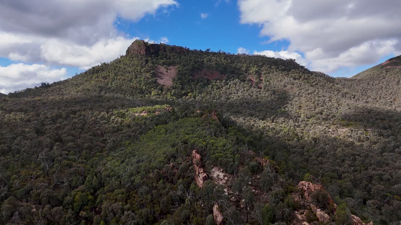 Drone camera glides steadily above dense forest toward a rugged mountain under partly cloudy skies, capturing Warrumbungle National Park’s dramatic landscape in natural daylight
