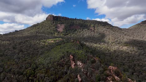 drone camera glides steadily above dense forest toward a rugged mountain under partly cloudy skies, capturing warrumbungle national park’s dramatic landscape in natural daylight