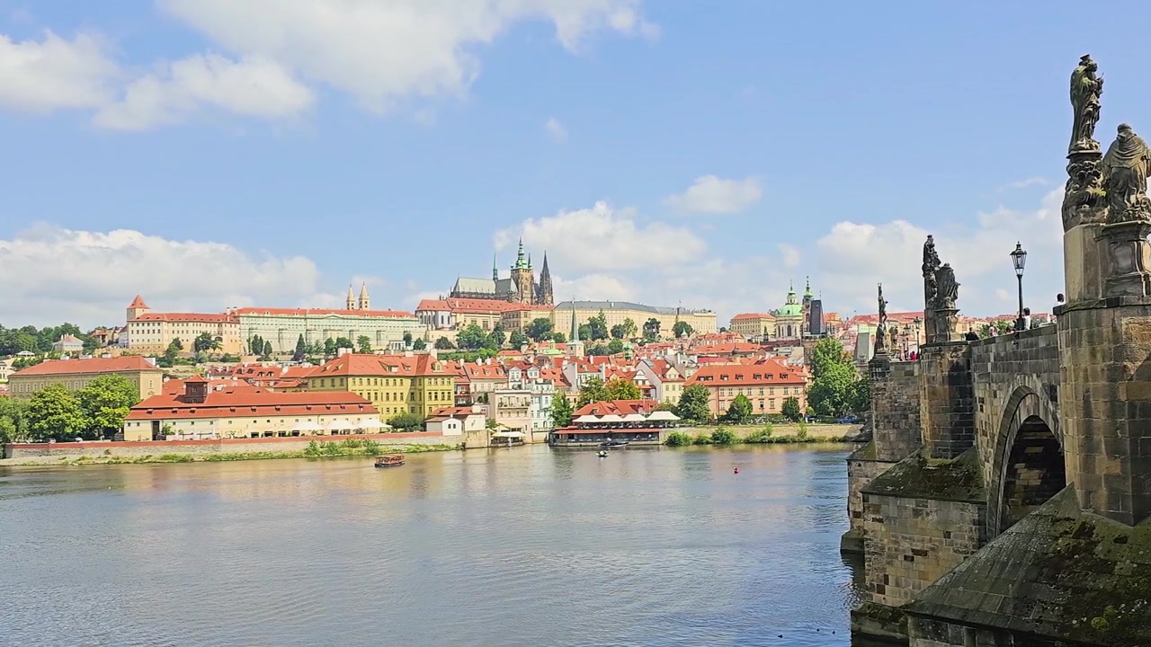 Paesaggio urbano soleggiato di Praga dal Ponte Carlo, che mostra archi in pietra, edifici lungo il fiume, tetti in tegole arancioni, il Castello di Praga, la Cattedrale di San Vito, il tranquillo fiume Moldava e un luminoso scenario di viaggio.