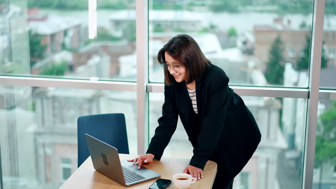 Dark-haired Caucasian woman stands leaning on the desk. Lady uses her laptop in the office with panoramic window. Top view.