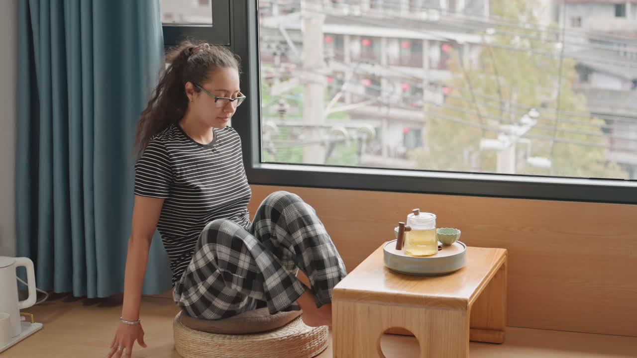 Ospite femminile in piedi vicino al finestrino dell'hotel che si gira verso il vassoio e la vista della città mentre pianifica l'itinerario. Stanza contemporanea con tavolino basso in legno, bollitore e snack sul vassoio, skyline urbano all'esterno, pensierosa.