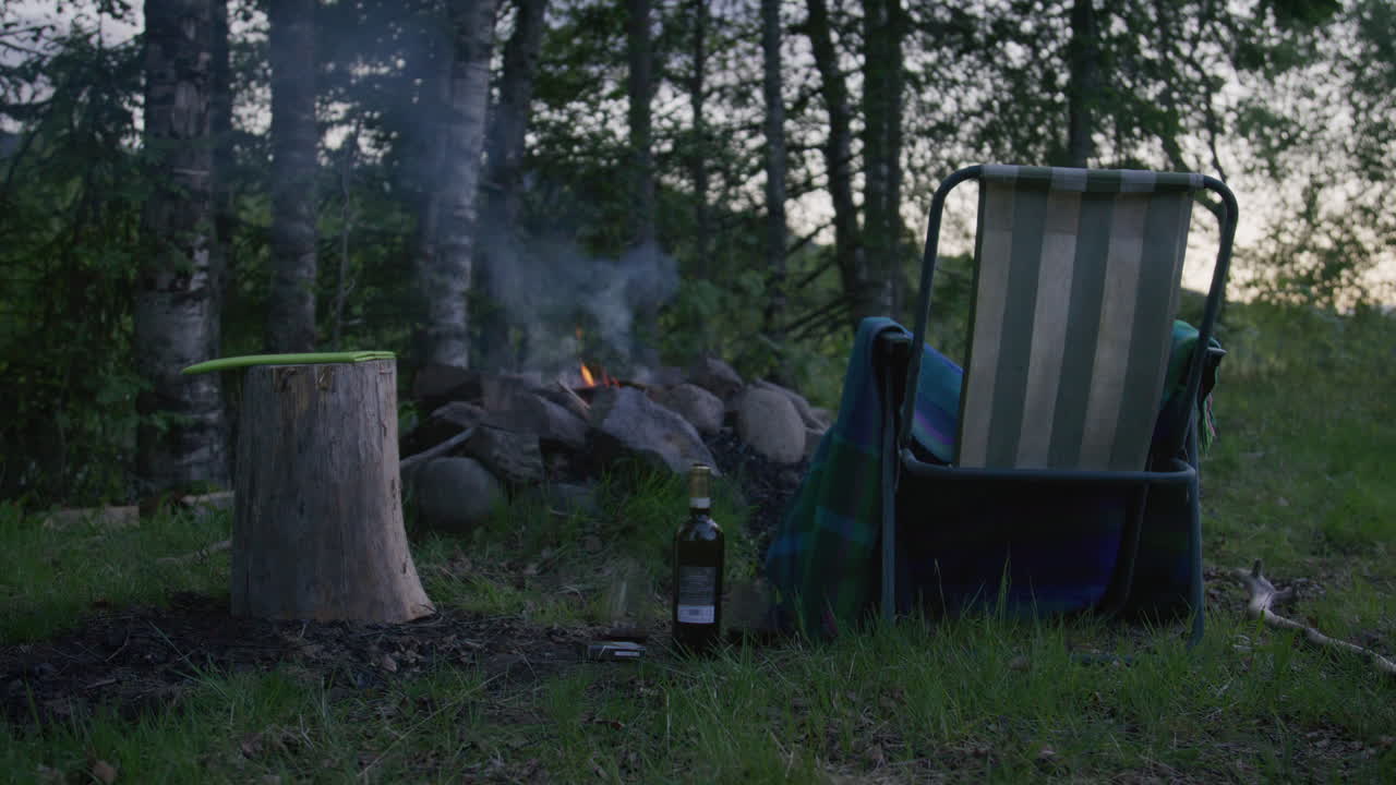 Campfire scene at forest edge with empty striped chair, chopping stump, and bottle on ground at dusk.