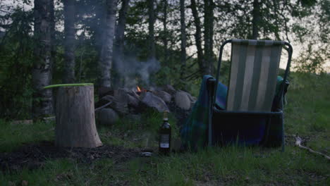 campfire scene at forest edge with empty striped chair, chopping stump, and bottle on ground at dusk.