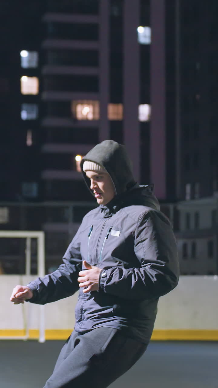 Side view of athlete in hoodie kicking soccer ball against wall under bright stadium lighting at night, urban cityscape with illuminated buildings and goalpost in background