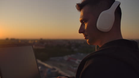 hipster man with a laptop on the edge of the roof. freelancer at work. wireless mobile internet. he works on the internet with headphones and listening music