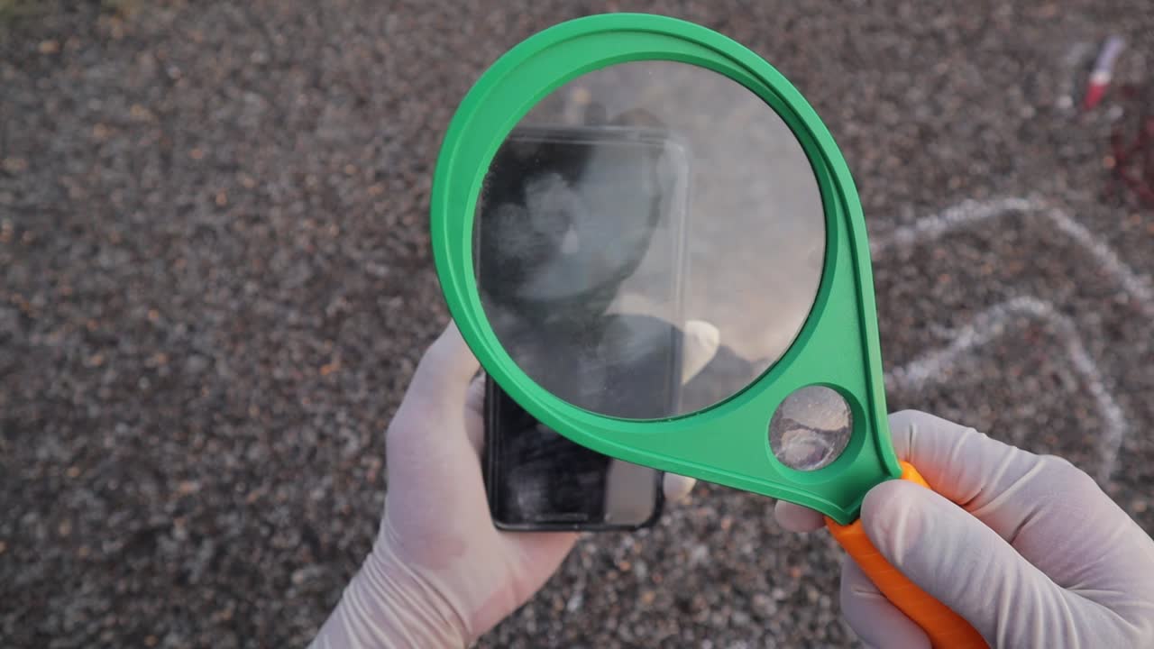 돋보기로 윤곽선이 그려진 분필 근처의 휴대폰을 확인하거나 검사하는 수사관