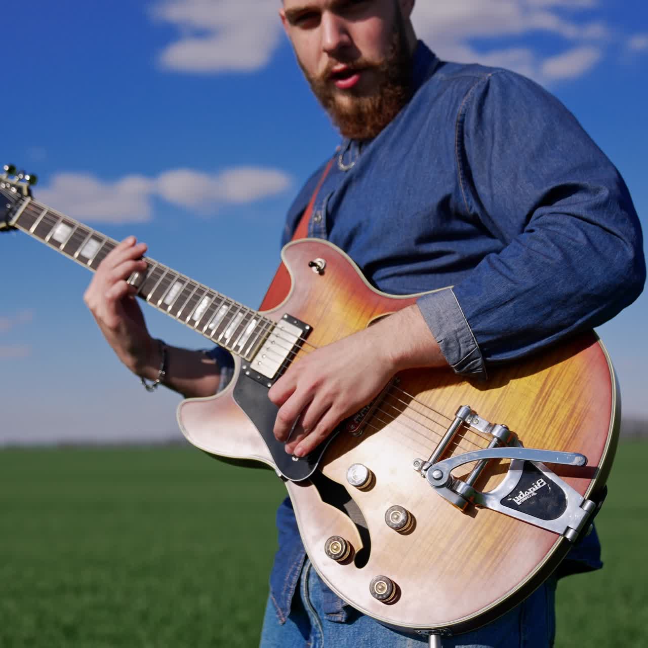 Handsome bearded guitarist in Jeanswear playing instrument and singing. Musician performing outdoors in the green field at backdrop of blue sky