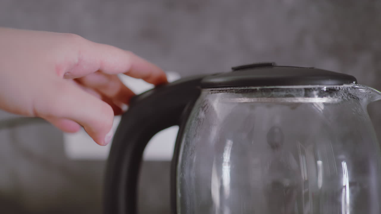 Close up of electric kettle filled with boiling water as person reaches to switch it off, with rising condensation on glass surface and hand partially visible in domestic kitchen environment