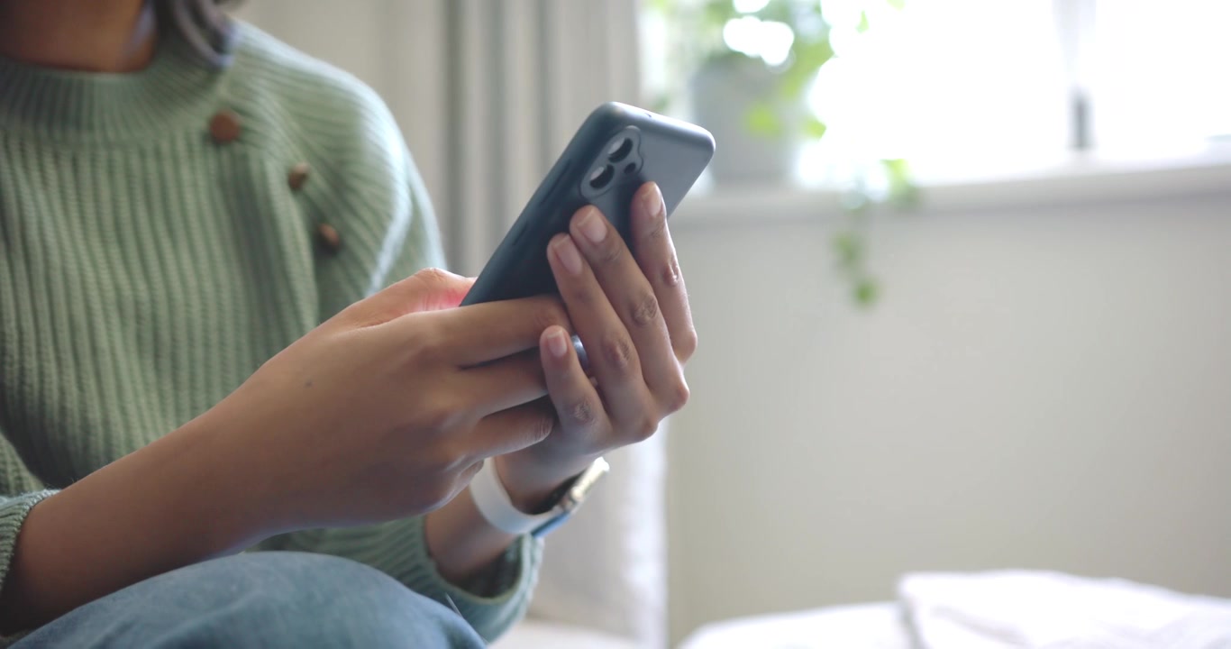 Afroameričanka na gauči u okna ťuká do smartphonu a reaguje na obsah, který čte na obrazovce.