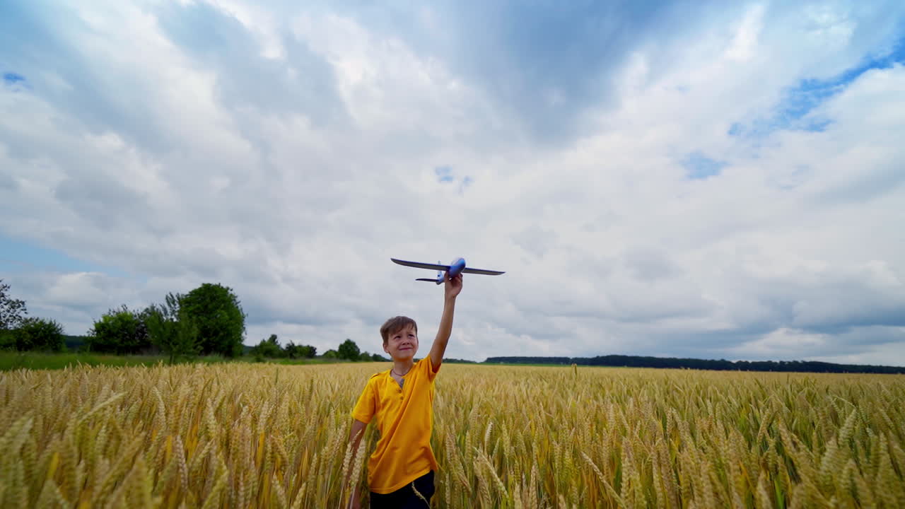 Little boy playing on field. Cute child in yellow t-shirt walks in agricultural land and holds the toy plane over his head. Front view.