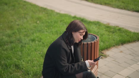 young woman in black coat and white sweater seated on park bench, focusing on her smartphone with a calm expression, surrounded by lush greenery