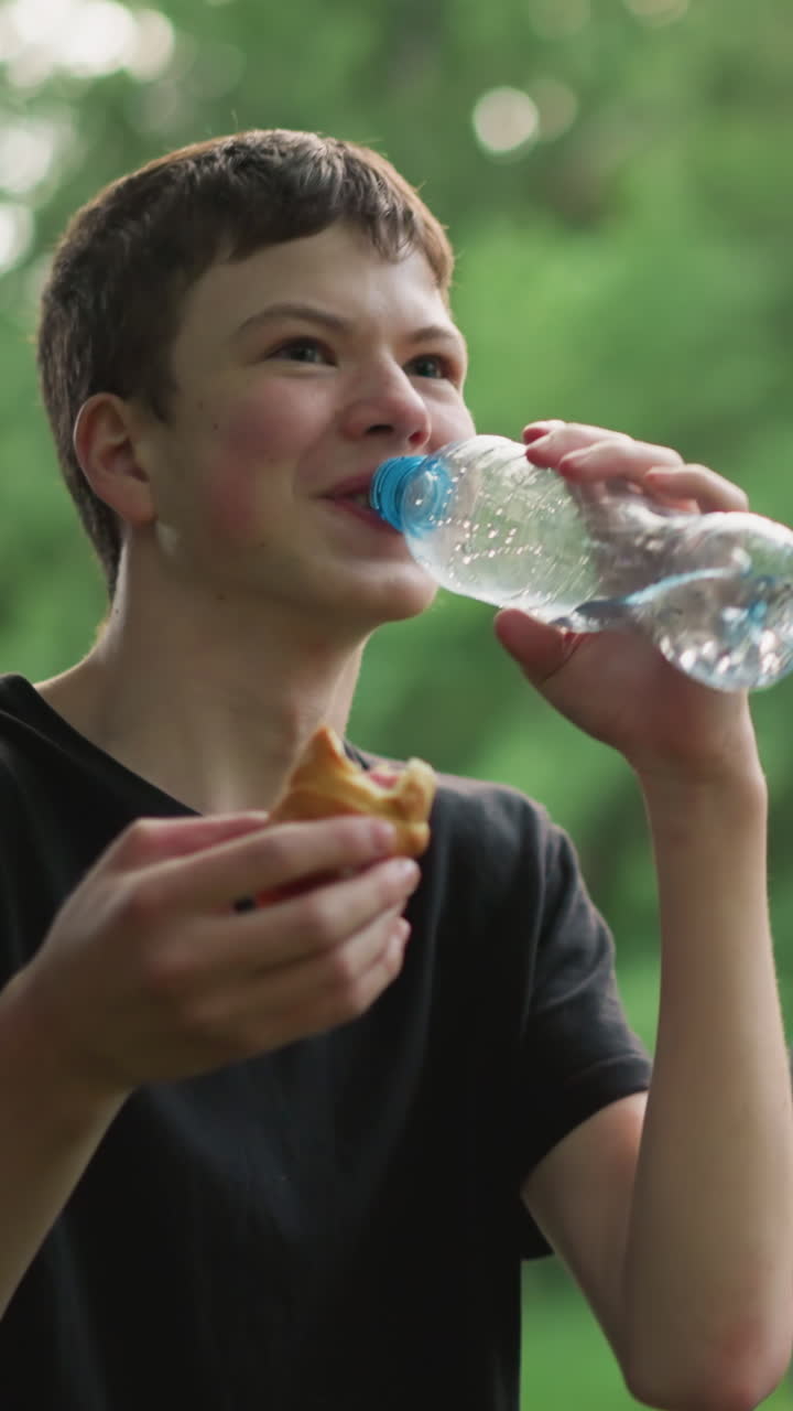 Young boy in black top bursts into laughter while drinking water and holding a snack, with his friend in the background cleaning his mouth and watching him laugh, surrounded by a greenery