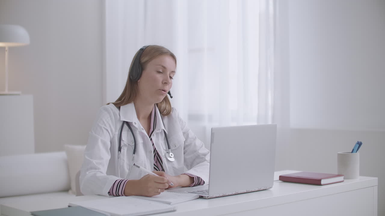 una joven terapeuta está consultando en línea preguntando al paciente y tomando notas en el cuaderno trabajando a distancia
