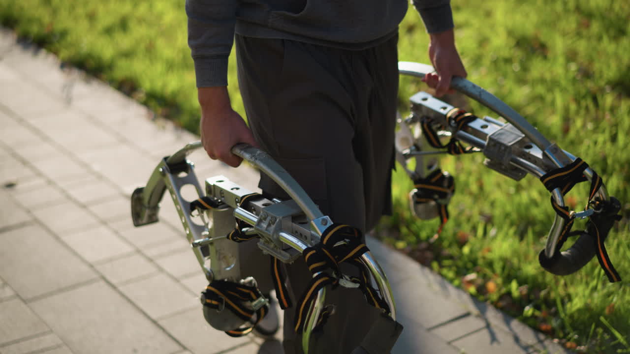 mitad inferior de la persona usando pilotes metálicos con soportes para los pies con correas negras, con pantalones sueltos de color gris oscuro y camisa de manga larga, caminando sobre una superficie pavimentada con fondo de hierba