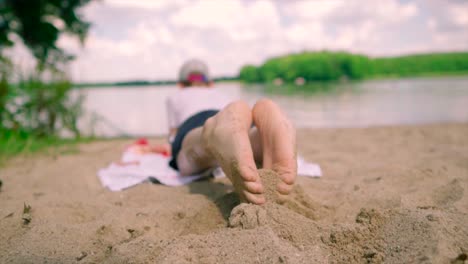 kid lifestyle: lay down on sand on a lakeshore in summertime playing with the sand with his legs